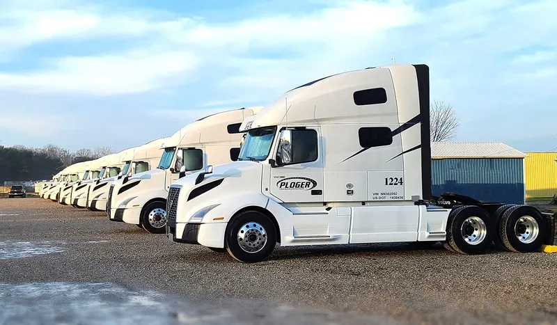 Row of white Ploger Transport semi trucks parked on gravel under a blue sky.