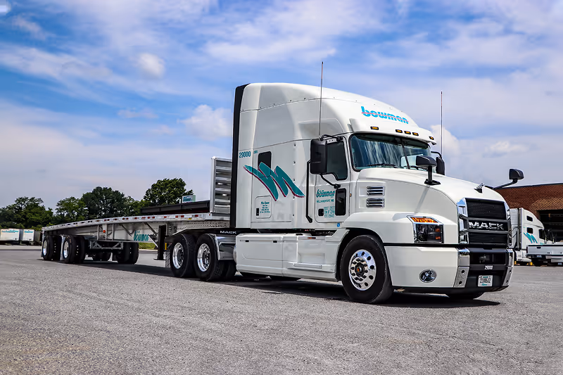 White Mack semi-truck with flatbed trailer parked on pavement under a partly cloudy sky.