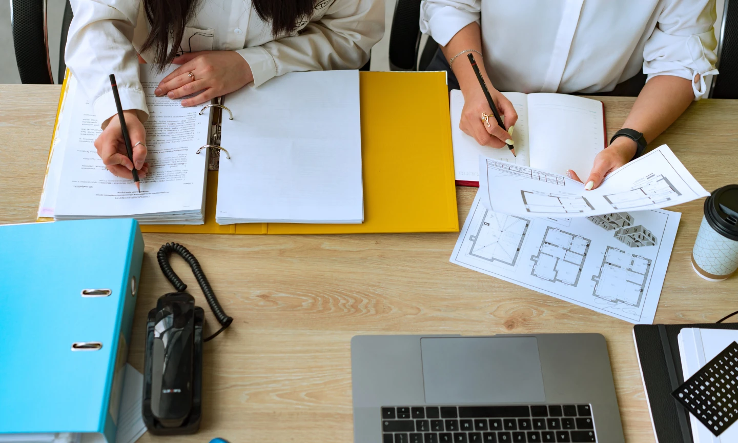 The main image of this comundo blog post about how you should check your a-rated buildings first shows two people working at a desk. We see it from above, and can only see their hands. On the table is a laptop, some paper and a large folder.