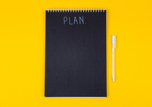 Wide view image of blank black spiral note pad and white marker with calligraphic inscription plan on yellow background