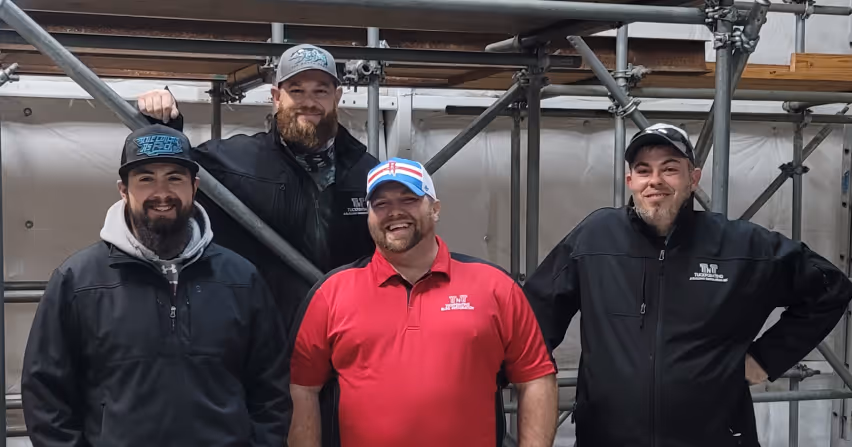 Four men smiling and posing together in front of metal scaffolding, three wearing black jackets and caps, one wearing a red polo shirt and a white and blue cap.