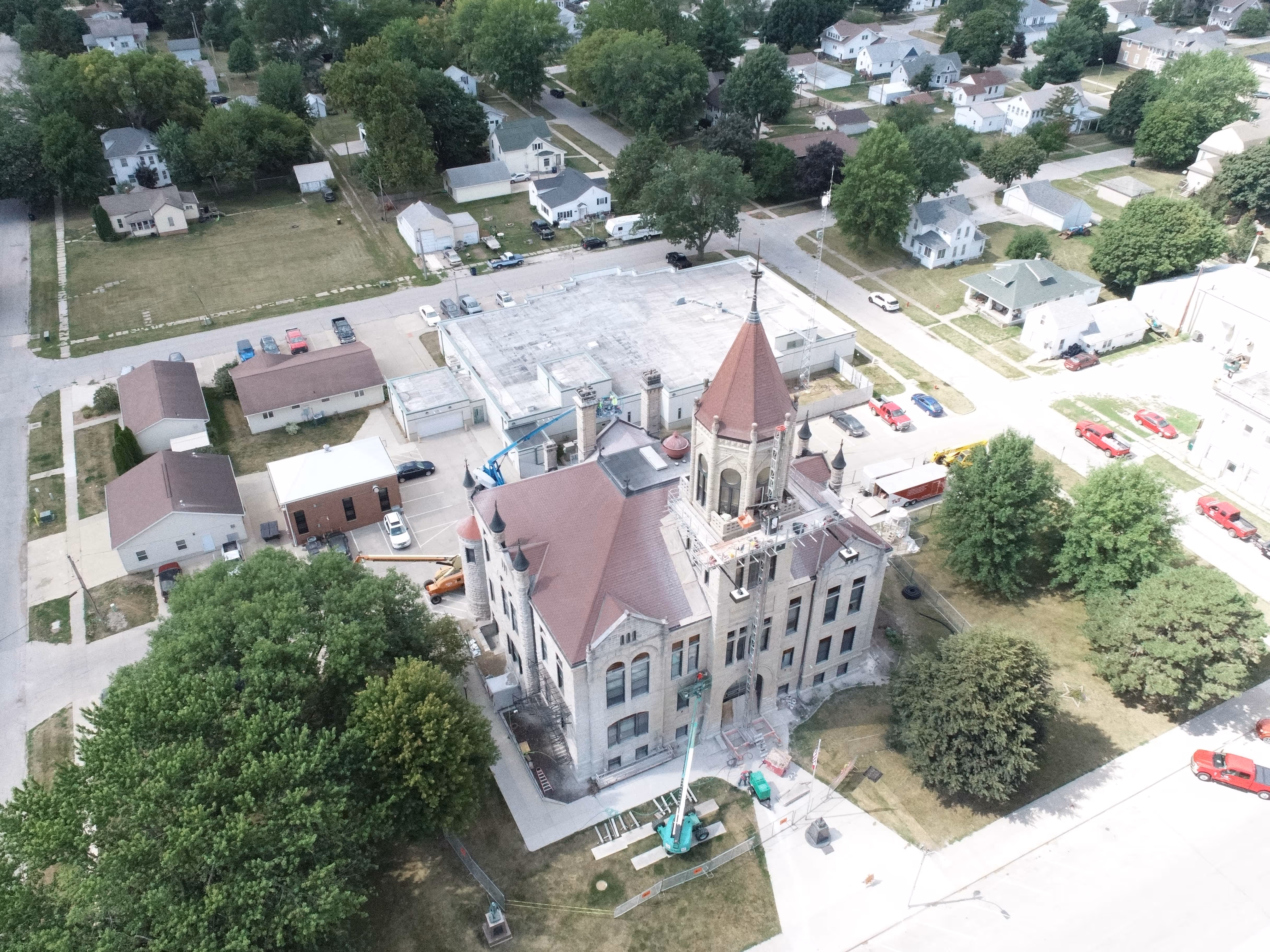 Aerial view of a historic stone building with a tall red-roofed central tower surrounded by trees and adjacent streets with parked vehicles.