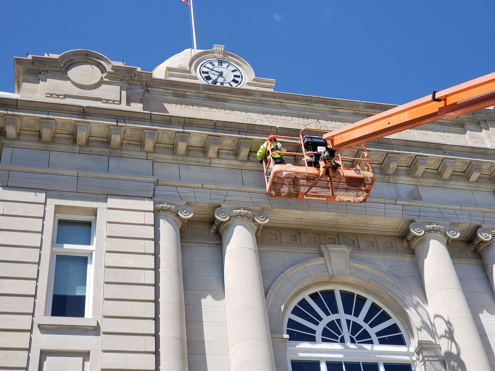 Worker in a lift basket performs maintenance near the clock on the classical facade of a stone building under a clear blue sky.