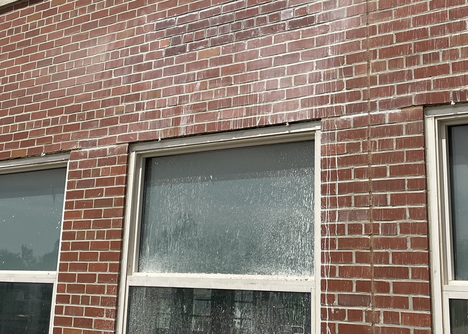 Exterior view of a red brick building featuring a large window. The horizontal row of bricks directly above the window frame is visibly bowing downward in the center, resting unevenly on the window casing. White streaks of residue cover the brickwork and the window glass.