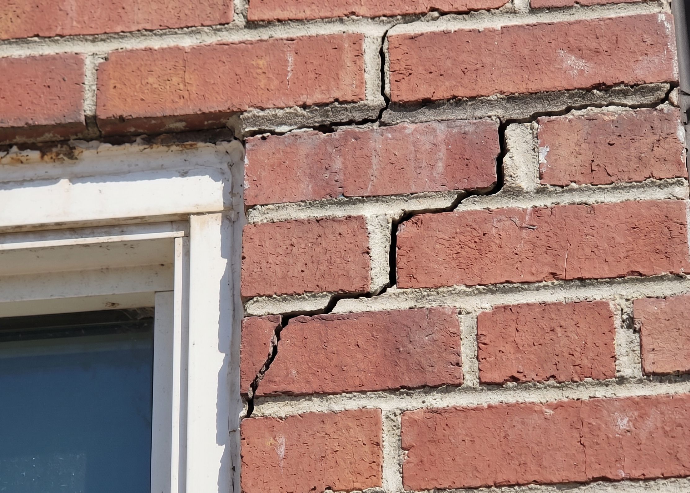 A close-up view of a red brick wall at the top right corner of a white window frame. A distinct stair-step crack zigzags diagonally upward and to the right through the mortar joints, originating near the edge of the window.