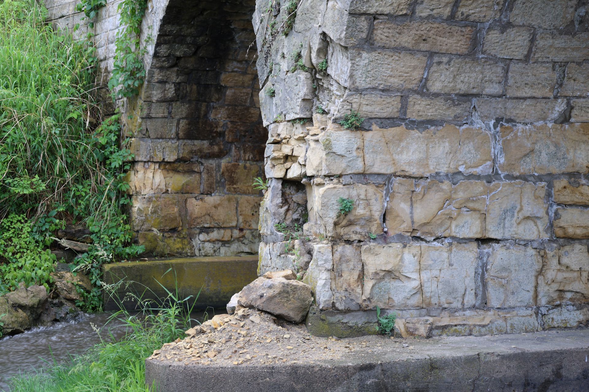 Eroded limestone at a pier of Ely's Stone Bridge at creek level, with fallen stone collected on the apron below.