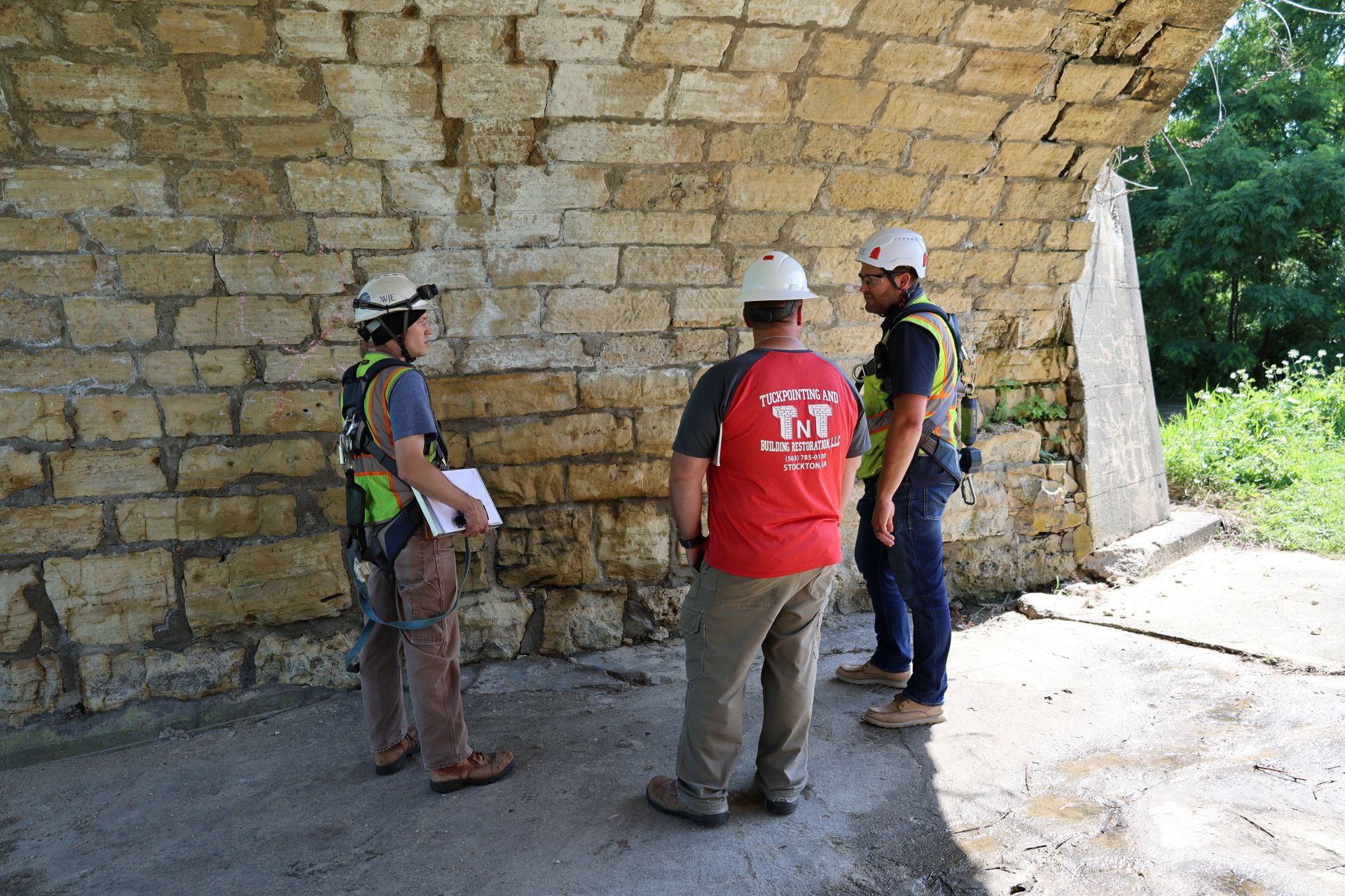WJE's engineer and architect reviewing the arch wall with TNT beneath the span, survey marks visible on the stone.