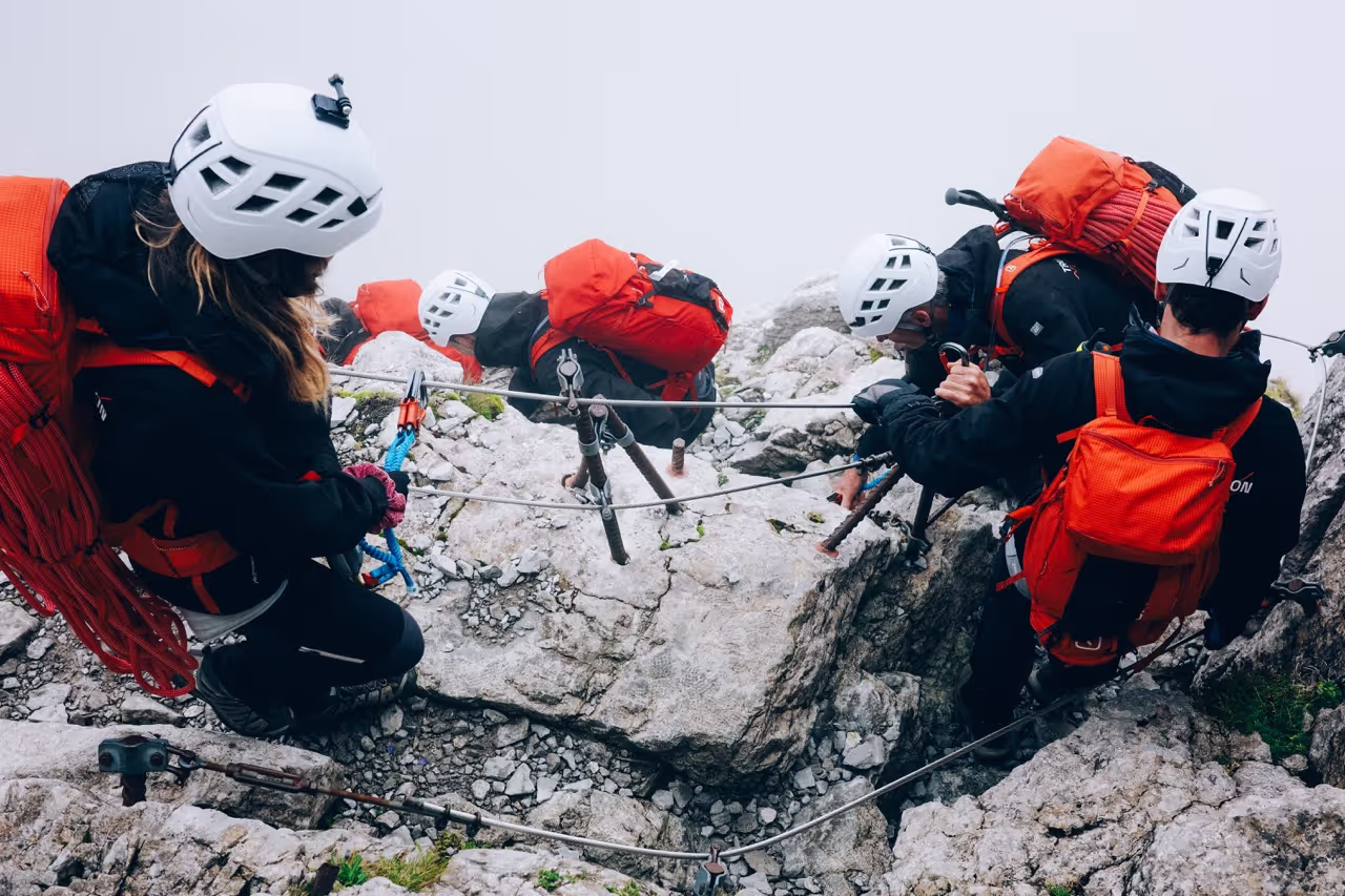 Vier bergbeklimmers met witte helmen en rode rugzakken klimmen een rotsachtige bergwand, gezekerd aan touwen.