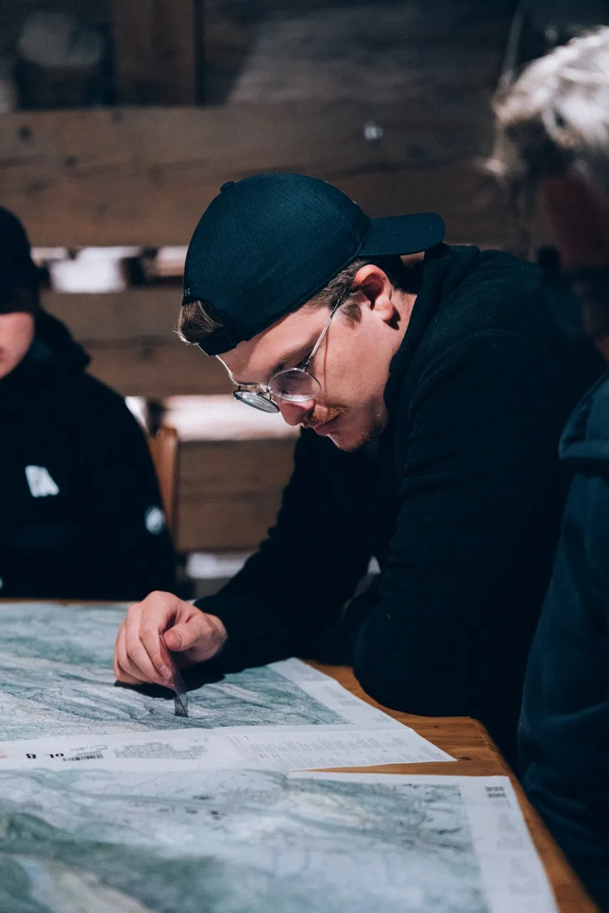 Man met pet en bril onderzoekt geconcentreerd een kaart op een houten tafel binnenshuis.