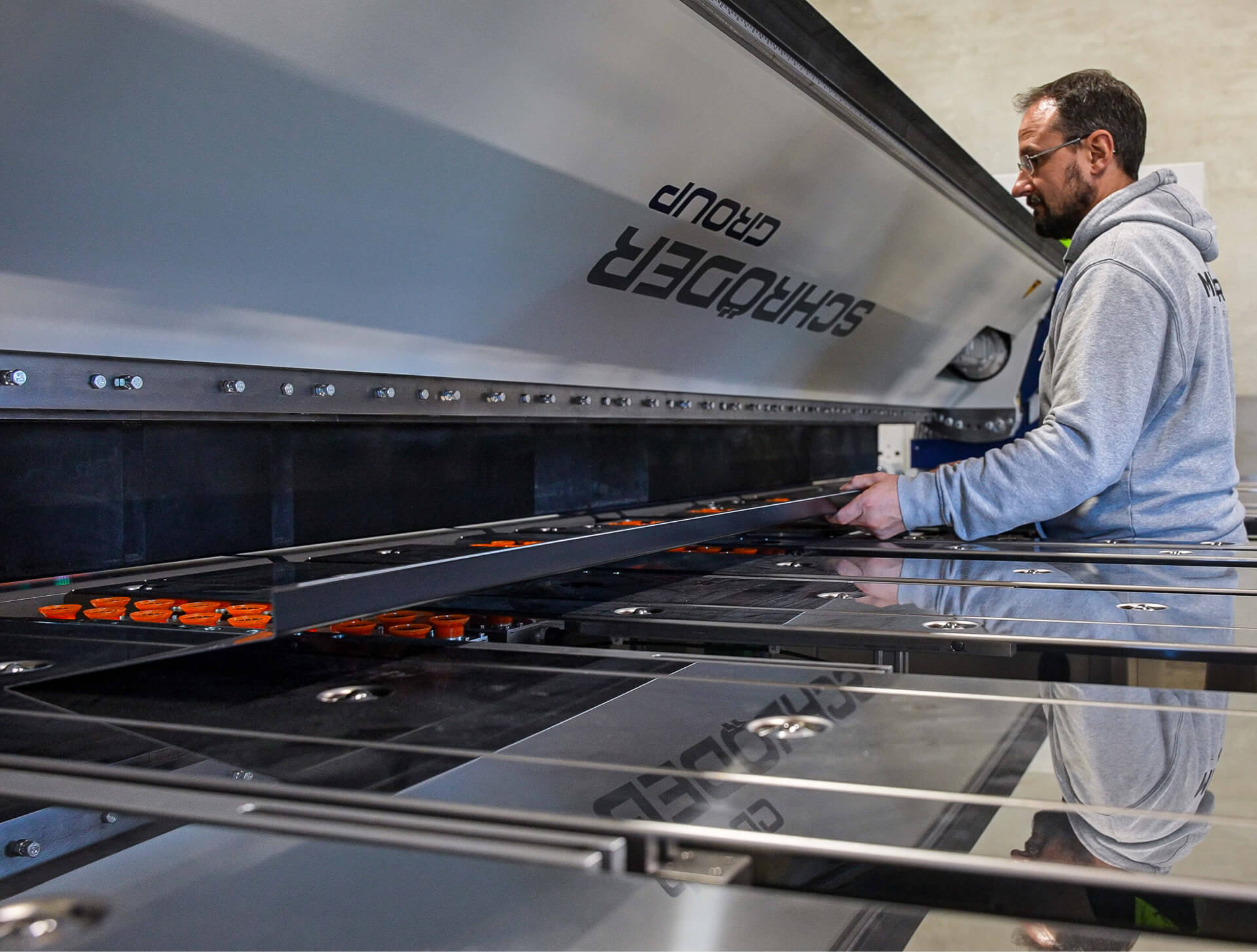 Man in a gray hoodie operating a Schröder Group industrial machine while holding a metal sheet.