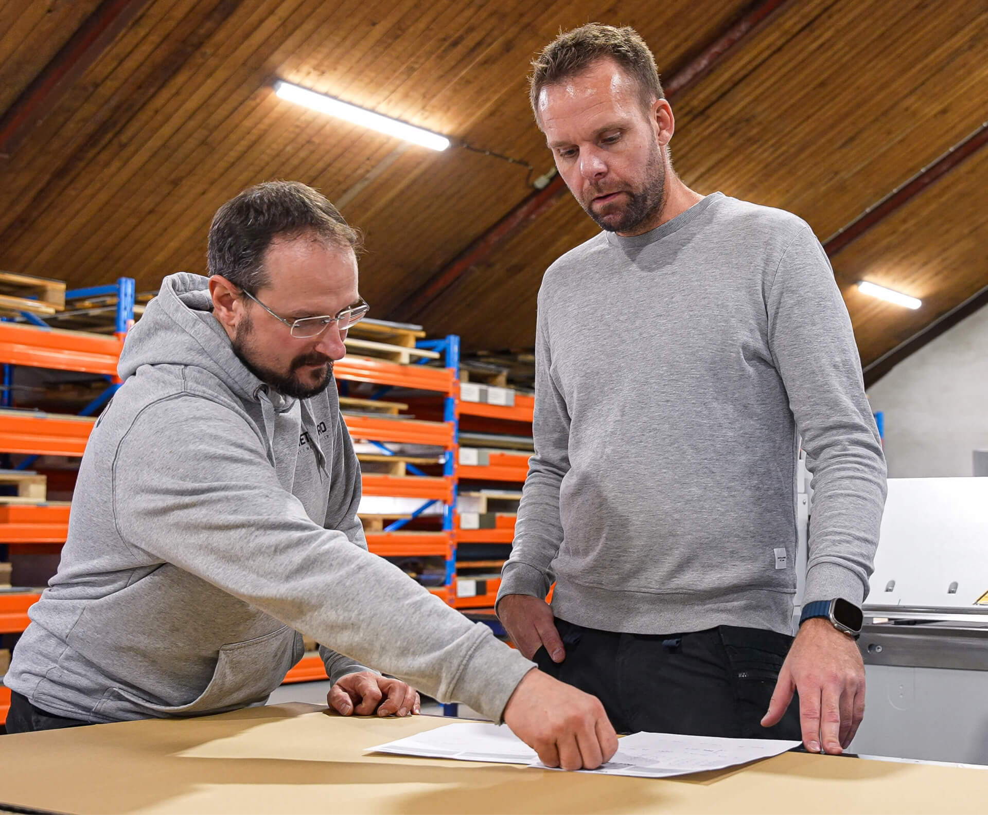 Two men in a workshop reviewing documents on a table with blue and orange shelving in the background.