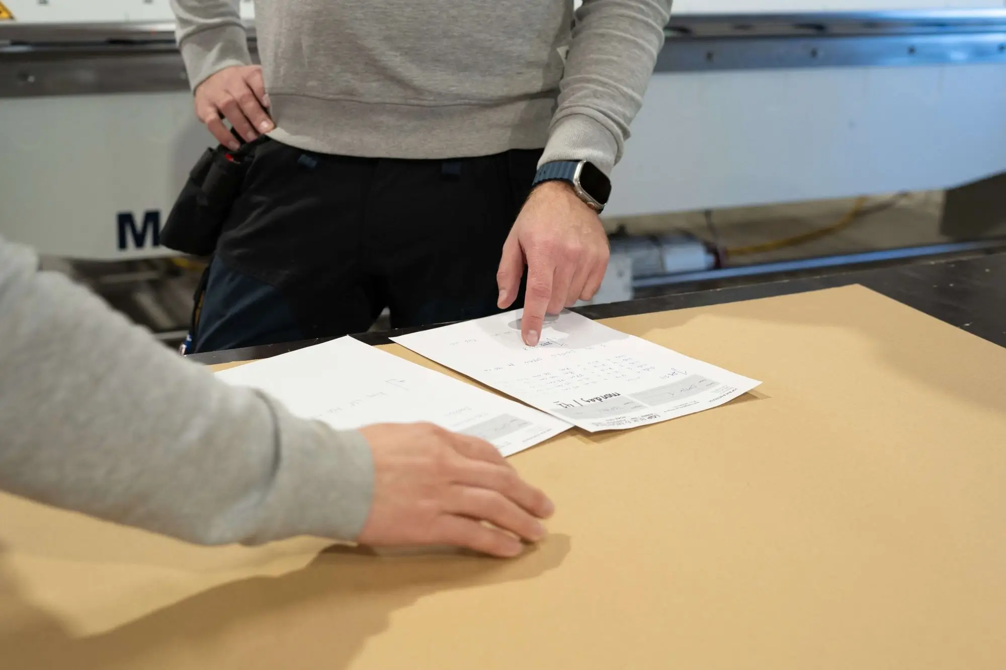 Two people reviewing and pointing at documents on a table in a workshop or industrial setting.