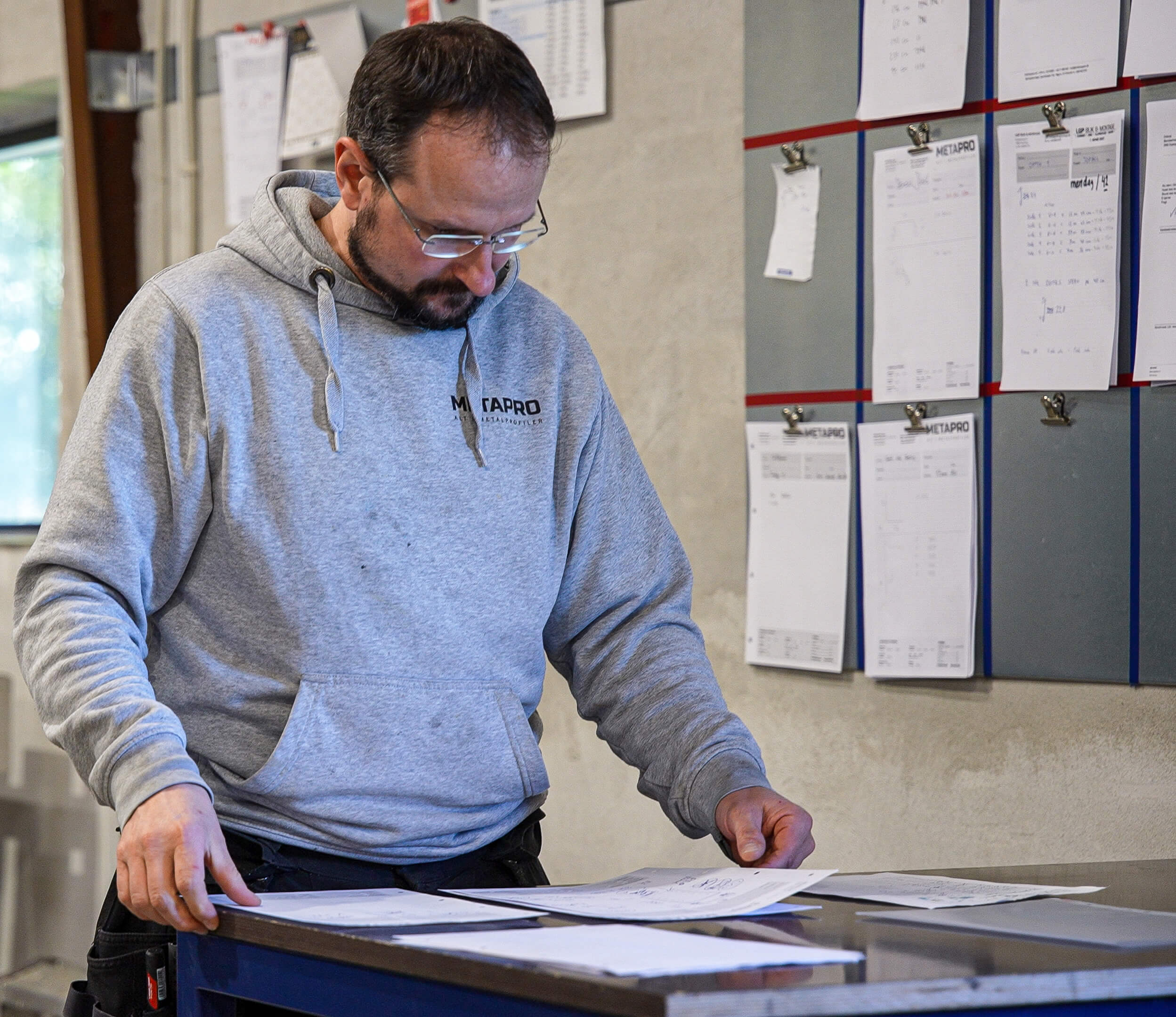 Man in gray Metapro hoodie reviewing papers on a desk with a bulletin board of pinned documents in the background.