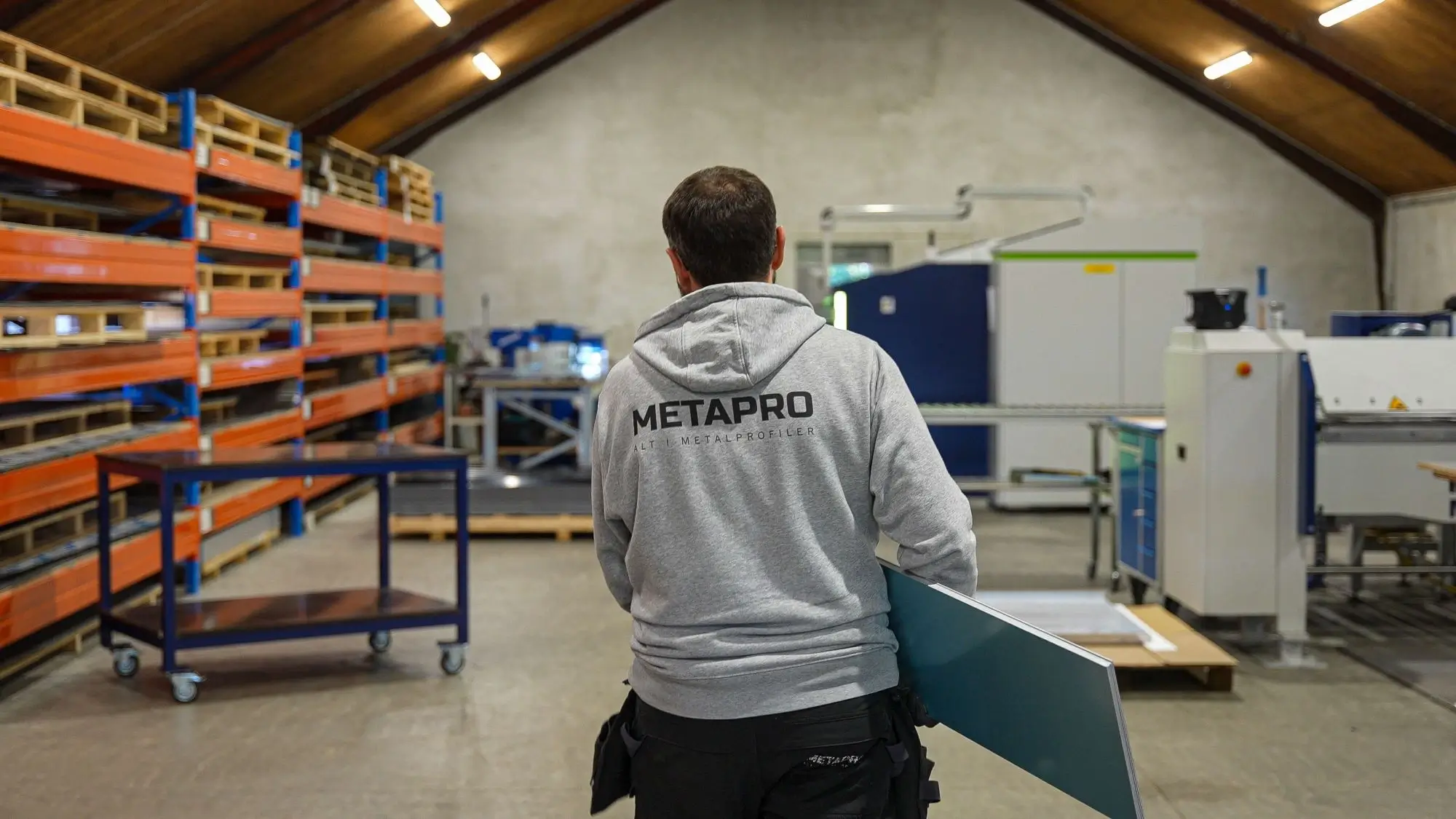 Worker wearing a gray METAPRO hoodie carrying a metal sheet in an industrial warehouse with shelves and machinery.