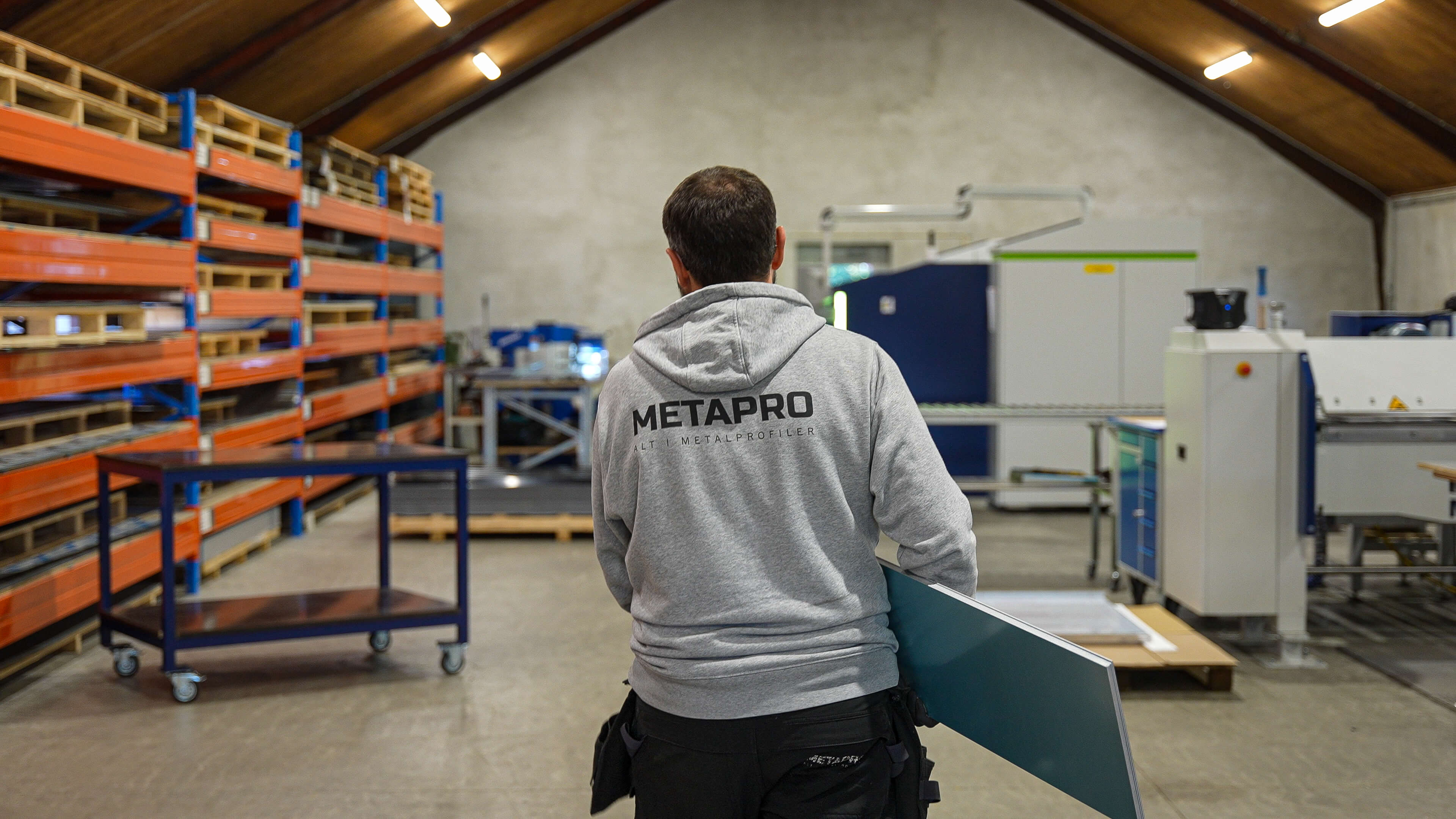 Worker wearing a gray METAPRO hoodie carrying a metal sheet in an industrial warehouse with shelves and machinery.
