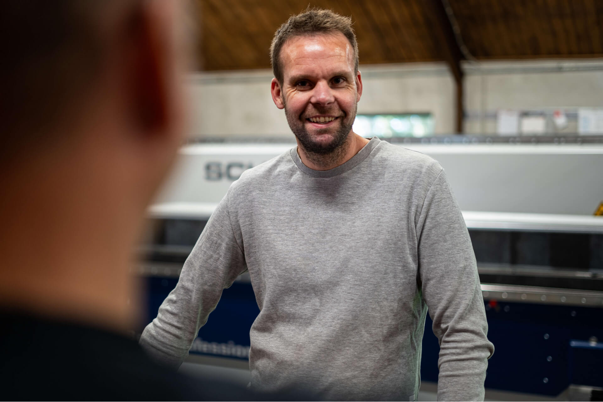 Smiling man in a gray sweater indoors, engaged in conversation with another person whose blurred head is in the foreground.