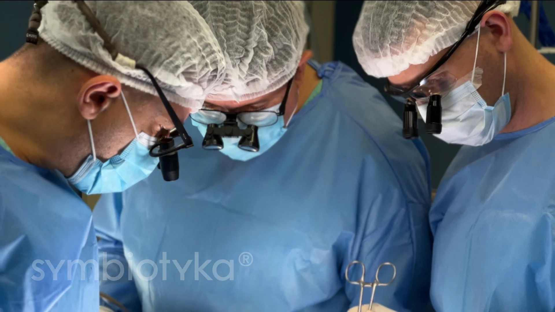 Three surgeons in blue scrubs, hair covers, and masks performing a surgery while wearing magnifying glasses.