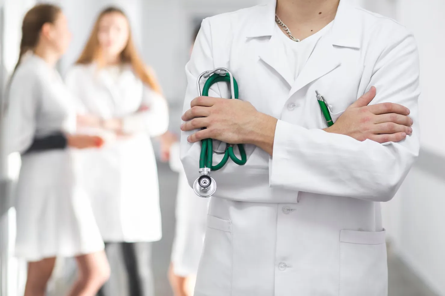 Doctor in white coat holding green stethoscope with blurred medical staff in the background.