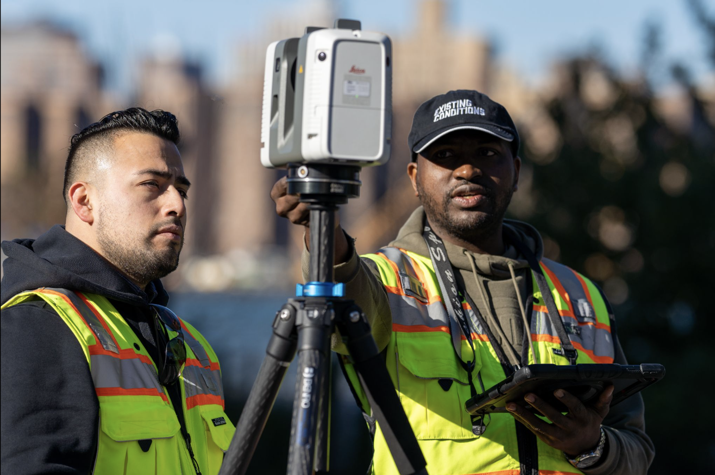 Two men in safety vests stand beside a tripod, engaged in a discussion or preparing for a task.