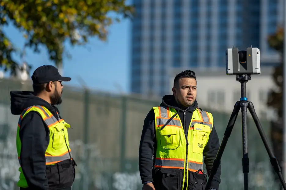 Two Project Managers in high-visibility, yellow vests standing outdoors near a tripod-mounted Leica RTC360 laser scanner, with a fence and tall building in the background.