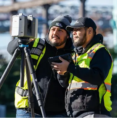Two Existing Conditions Project Managers wearing black hats, black sweatshirts, and bright yellow vests operating a Leica Laser Scanner on a tripod. One of them is holding an iPad in a black case. 
