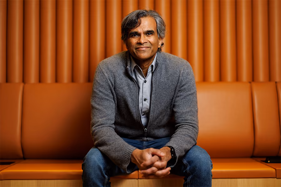 A portrait of Sendhil Mullainathan, seated on a bench leaning forward, hands clasped, partially smiling.
