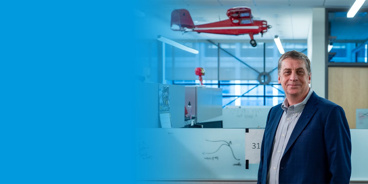 an image of Jonathan How, the professor of Aeronautics and Astronautics and the Director of the Aerospace Controls Laboratory at MIT standing in an office with a red model airplane hanging behind him from the ceiling above