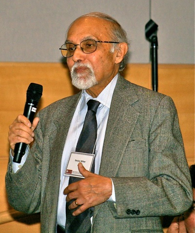 Closeup of Sanjoy Mitter, an older gentleman, speaking into a microphone in an indoor space