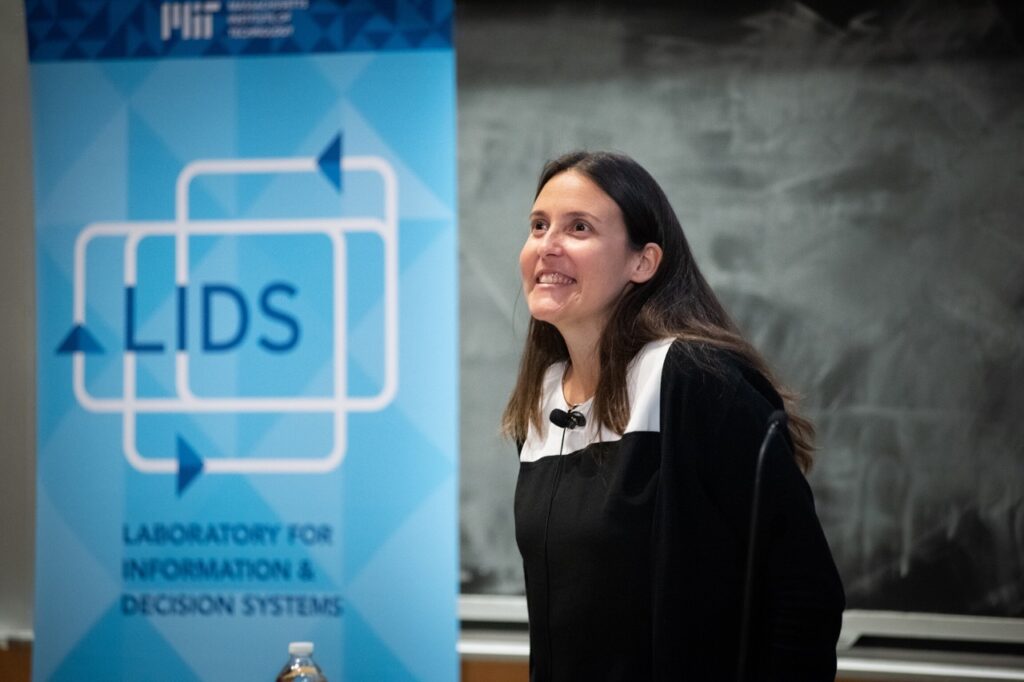 A photo of Caroline Uhler standing in front of a black chalkboard with a blue Laboratory for Information and Decision Systems banner to her right
