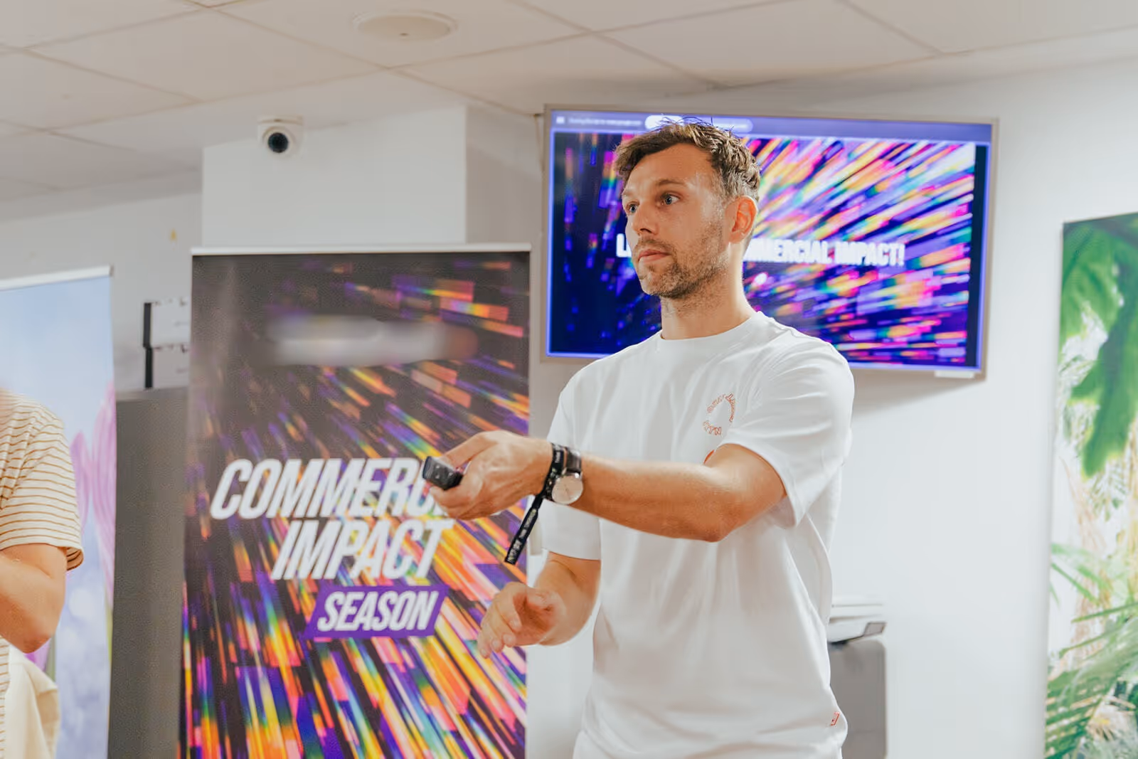 Man in white t-shirt giving a presentation in front of a colorful 'Commercial Impact Season' banner and screen.