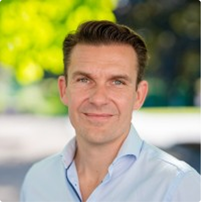 Man with short brown hair wearing a light blue shirt, smiling in an outdoor setting with blurred green and yellow foliage background.