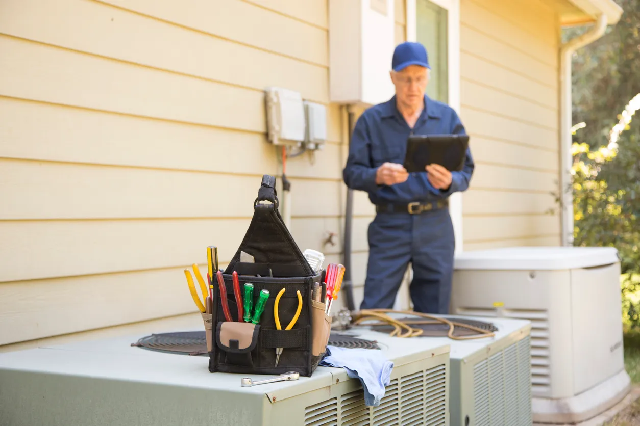 Technician in blue uniform with tablet beside outdoor air conditioner topped with tool bag and cables