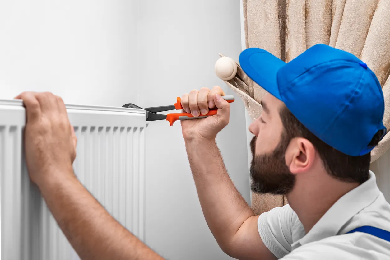 Person in blue cap and white shirt using wrench to fix radiator near beige curtain
