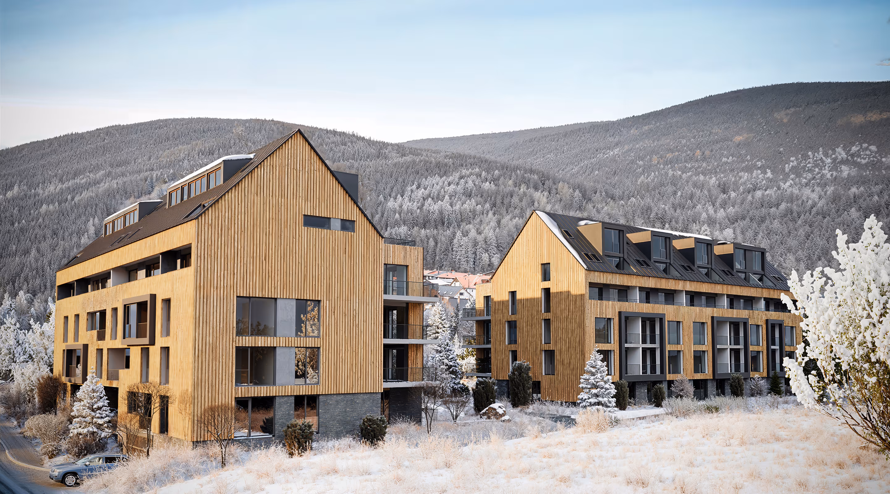 Dva moderní dřevěné bytové domy v zasněžené krajině s lesem a kopci v pozadí.