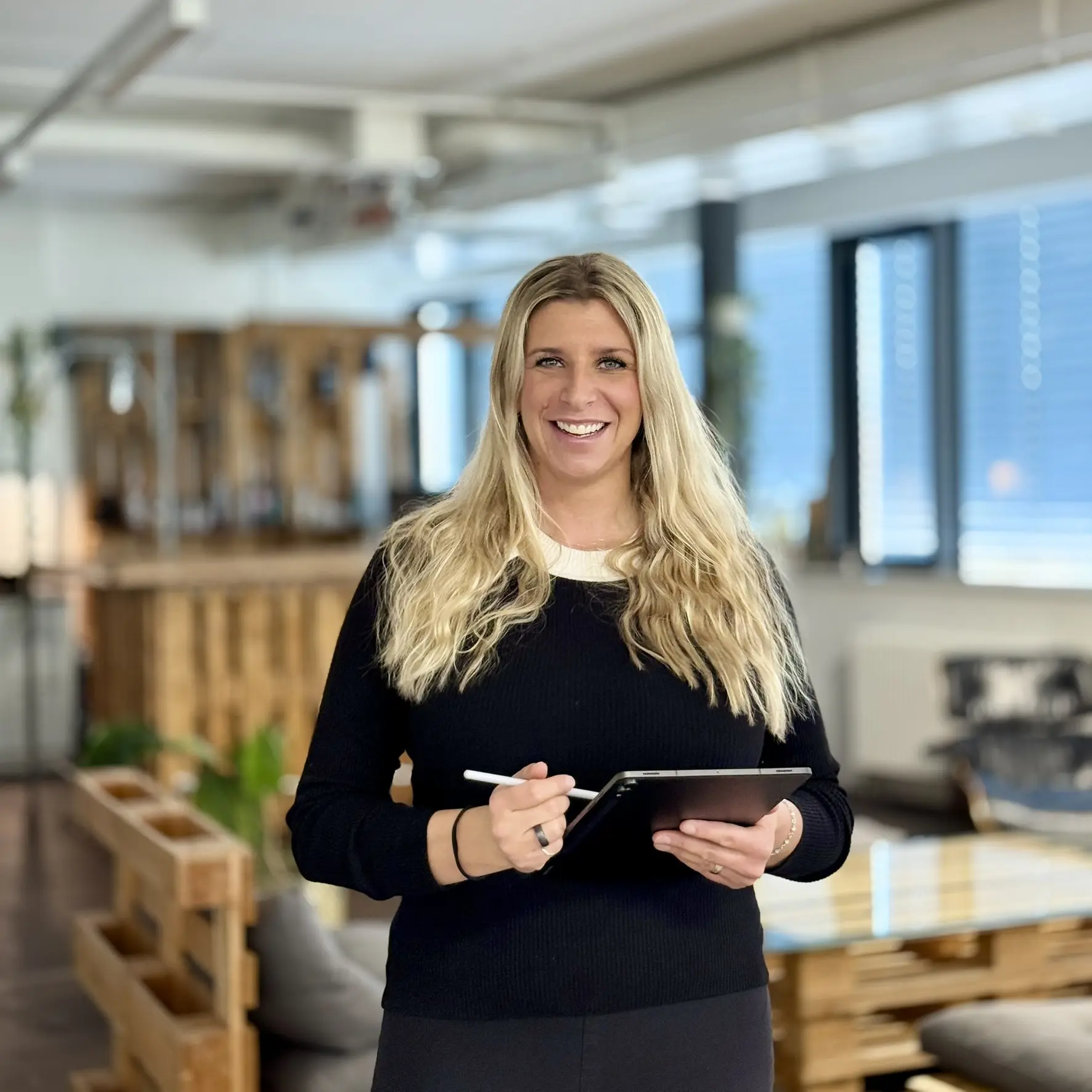Portrait Sarah Künzli with blonde ponytail, black top with button placket, in the background a shelf with books and plants.