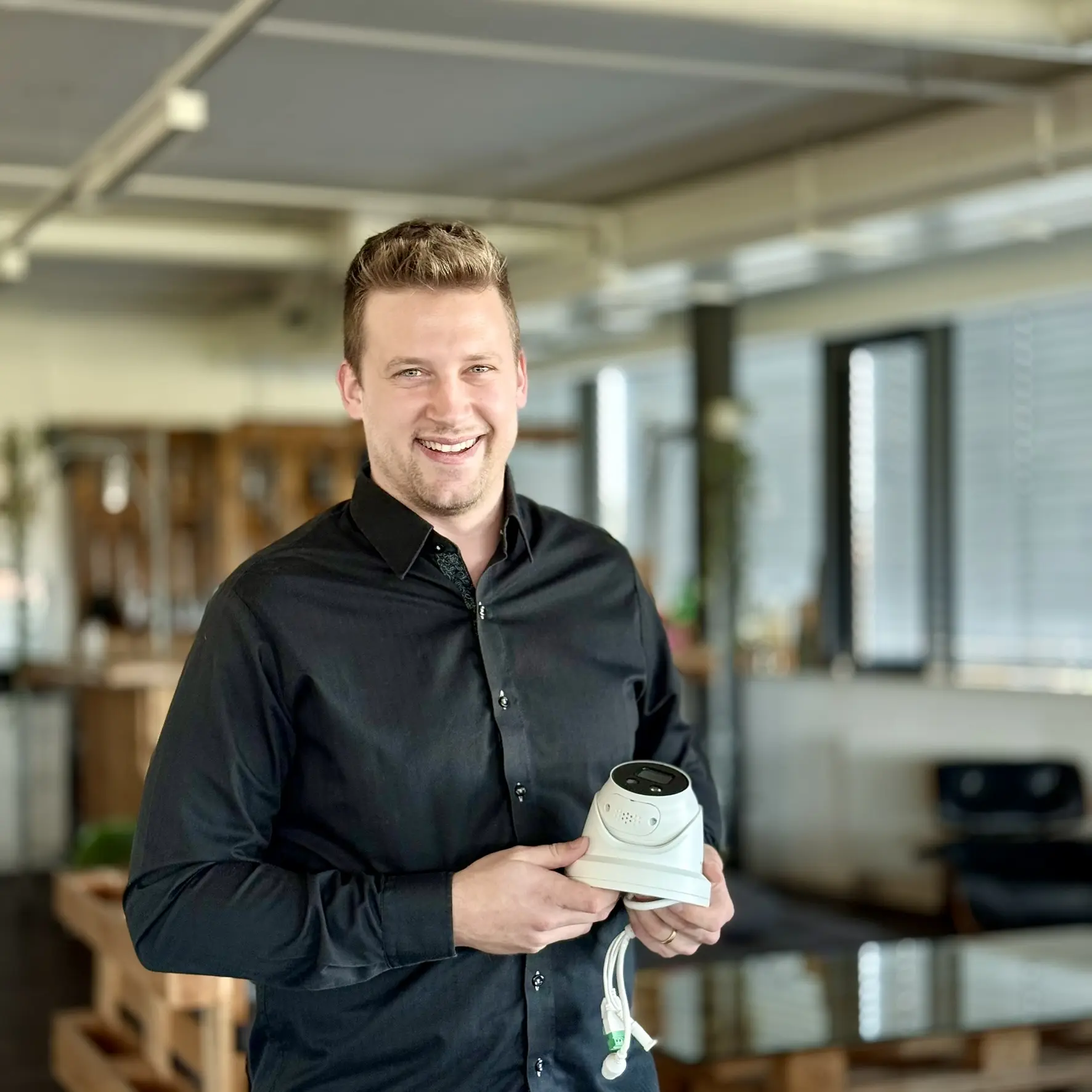 Portrait of Jonas Schmid with short, light brown hair and a black shirt, holding a surveillance camera, with a shelf with books and plants in the background.
