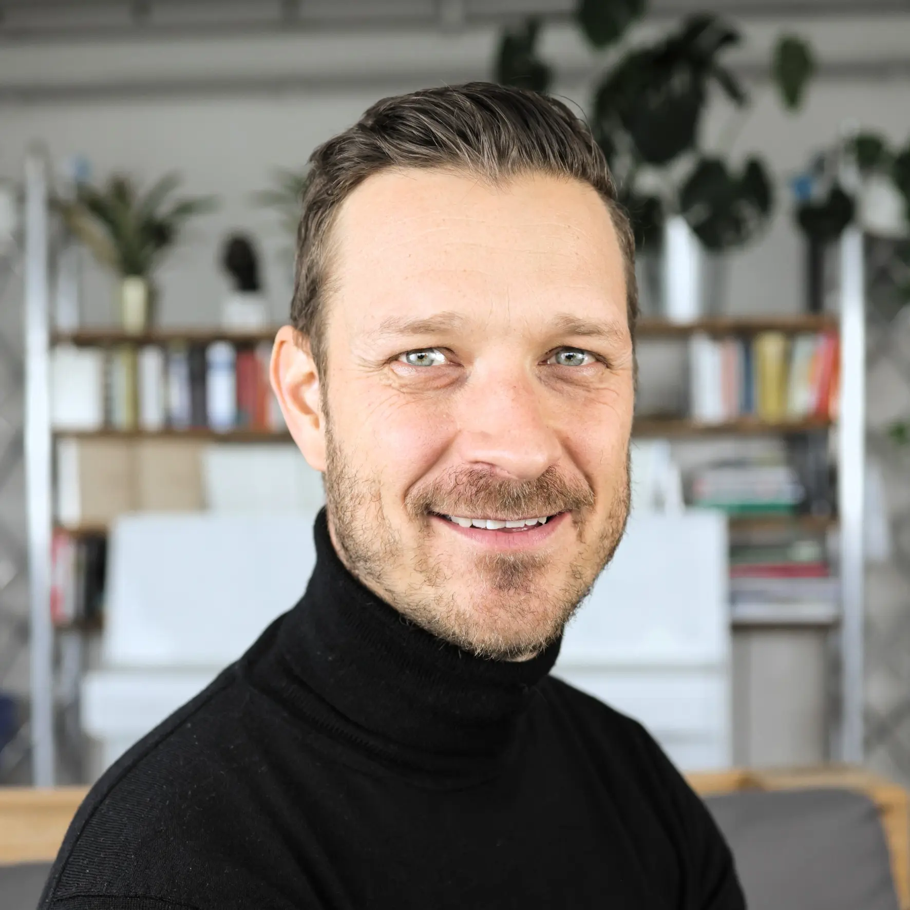 Picture Manuel Zingg, Managing Director homesecurity, with short brown hair and black turtleneck sweater, in the background a shelf with books and plants.