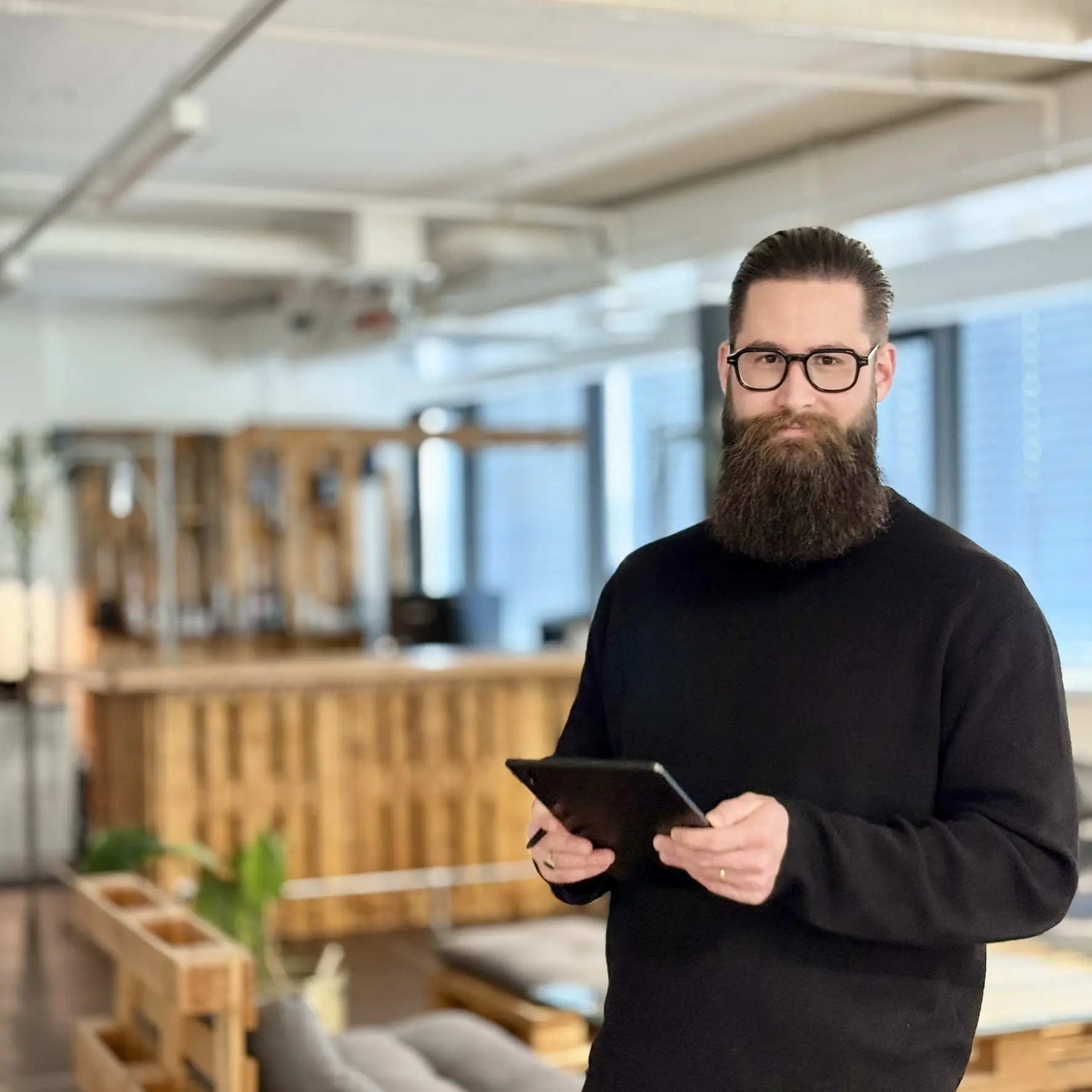 Portrait de Jonas Furer-Junker, cheveux bruns et longue barbe, tablette et stylo, souriant, regardant l'appareil photo, arrière-plan flou.