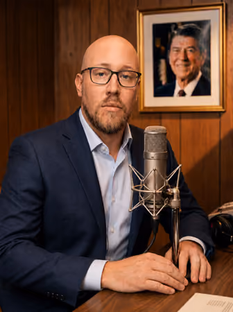 Radio host Walt Philips in a navy suit seated at a desk with a studio microphone and framed portrait in the background
