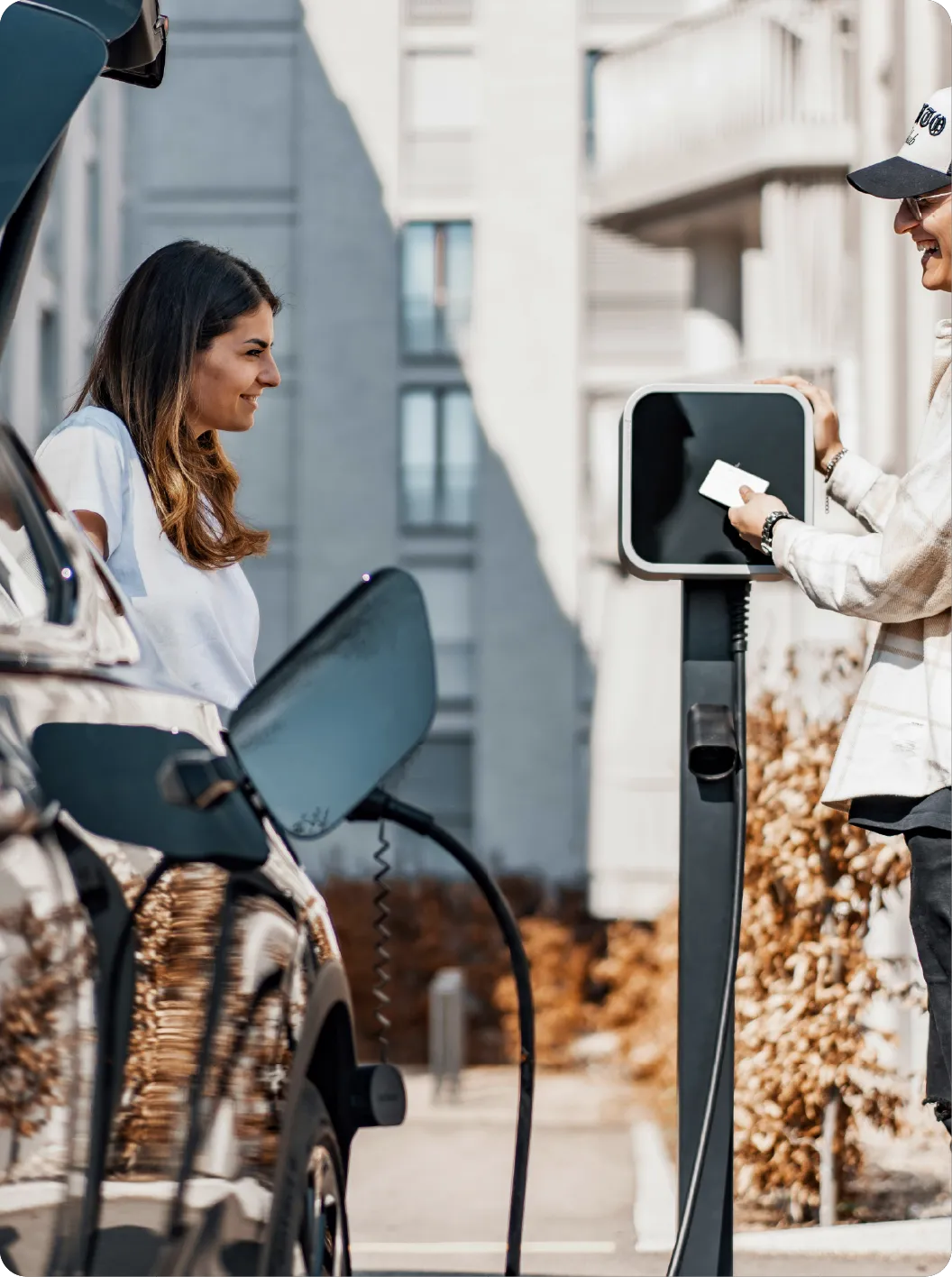 Une personne utilise une carte à une borne de recharge pour véhicule électrique tandis qu'une femme sourit à côté d'une voiture en cours de recharge.
