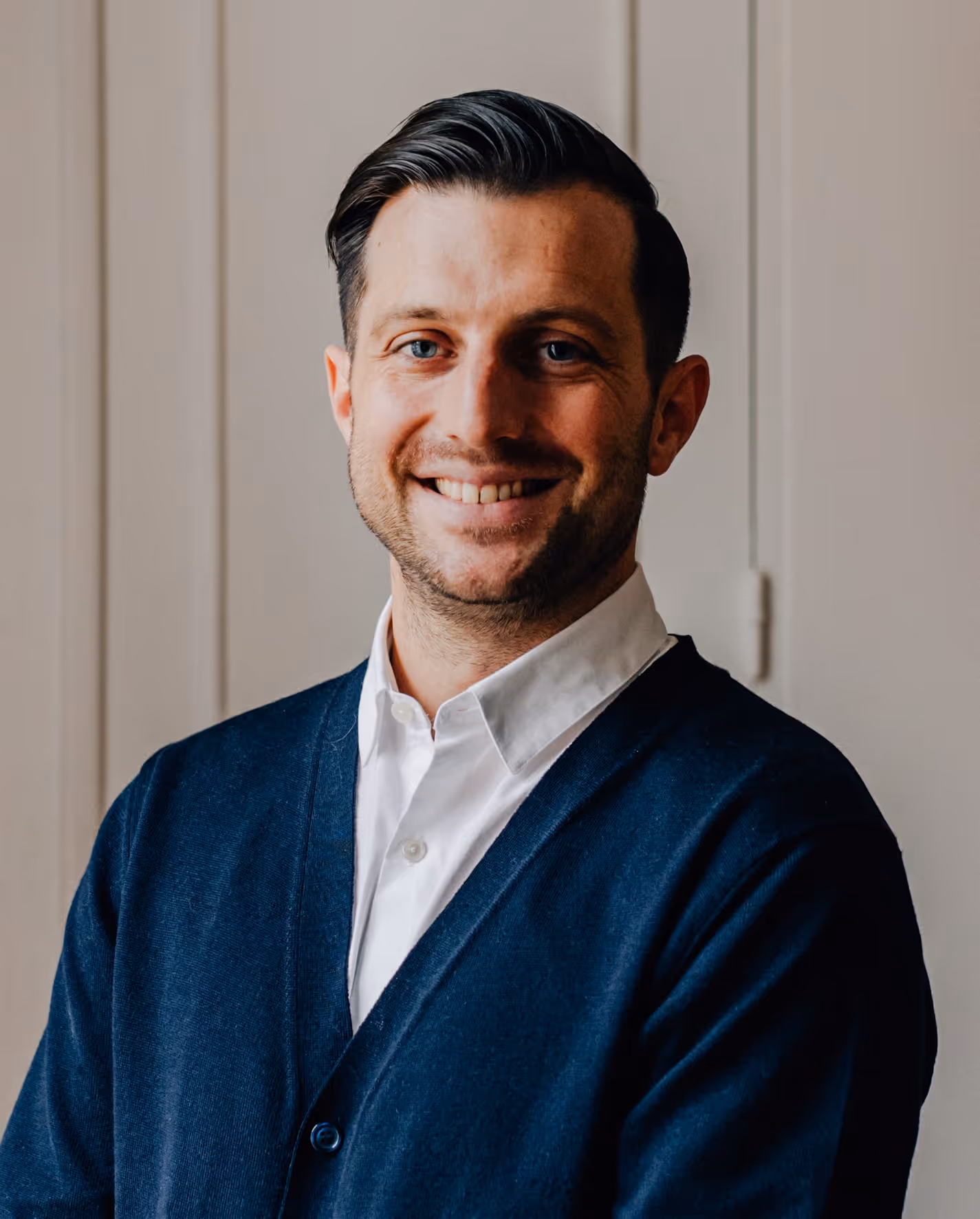 Homme souriant aux cheveux courts et foncés, vêtu d'une chemise blanche et d'un cardigan bleu marine, devant un fond clair.