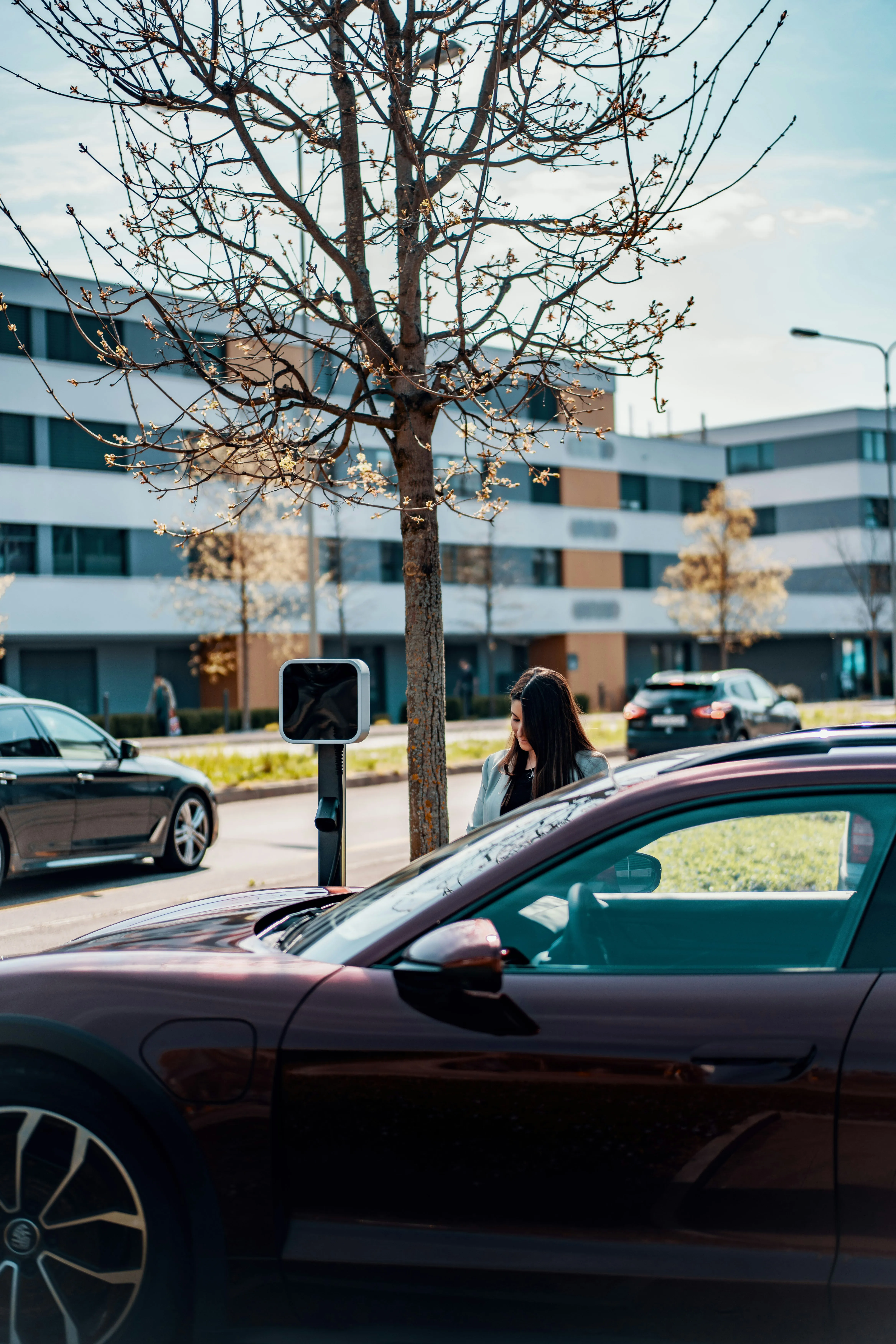 Woman standing next to an electric car charging station in an urban area with modern buildings in the background.