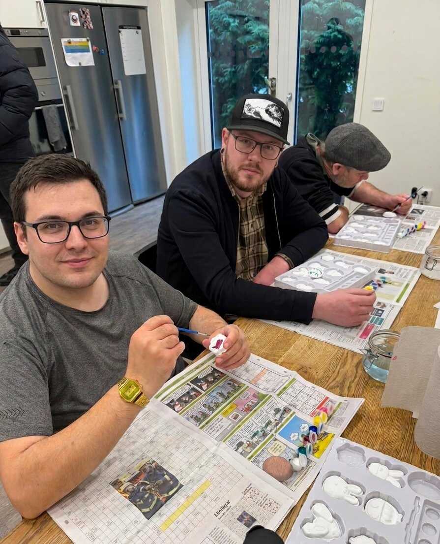 Tre personer sitter vid ett bord och målar små gipsfigurer med akrylfärg, med tidningar som underlag