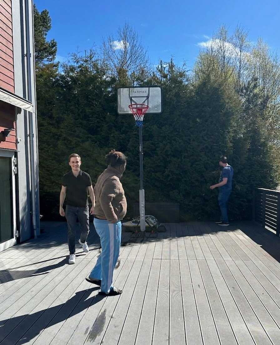Boende spelar basket på en uteplats i solen, med en portabel basketkorg och träd i bakgrunden