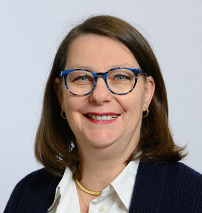Portrait professionnel d'une femme souriante, portant des lunettes à monture bleue, une chemise blanche et un gilet bleu marine, sur fond clair neutre.