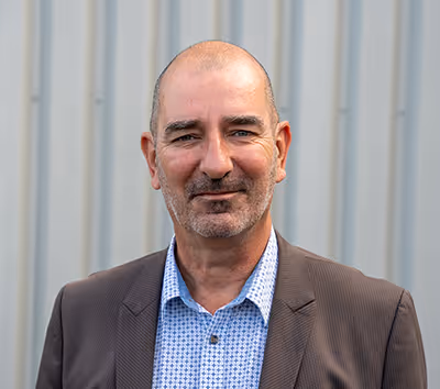 Portrait professionnel d’un homme en veste marron, chemise à carreaux, devant une paroi métallique claire.