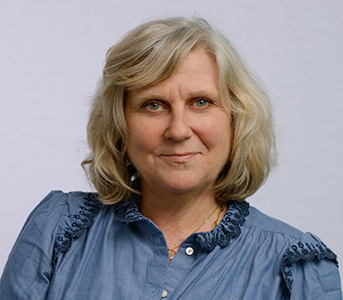 Portrait professionnel d'une femme aux cheveux gris-blonds, portant une chemise bleue à col brodé, sur fond uni clair