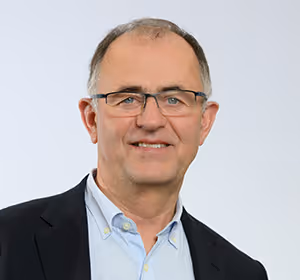 Portrait d’un homme avec lunettes, cheveux courts grisonnants, portant un blazer foncé et une chemise bleu clair, sur fond uni clair