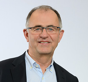 Portrait d’un homme avec lunettes, cheveux courts grisonnants, portant un blazer foncé et une chemise bleu clair, sur fond uni clair