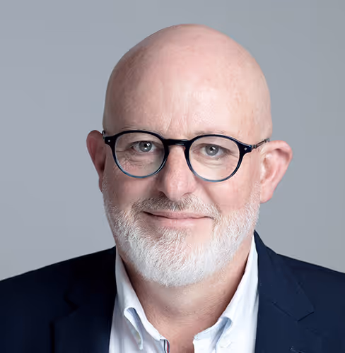 Portrait professionnel d’un homme chauve à lunettes rondes et barbe blanche, en costume bleu et chemise claire, sur fond gris